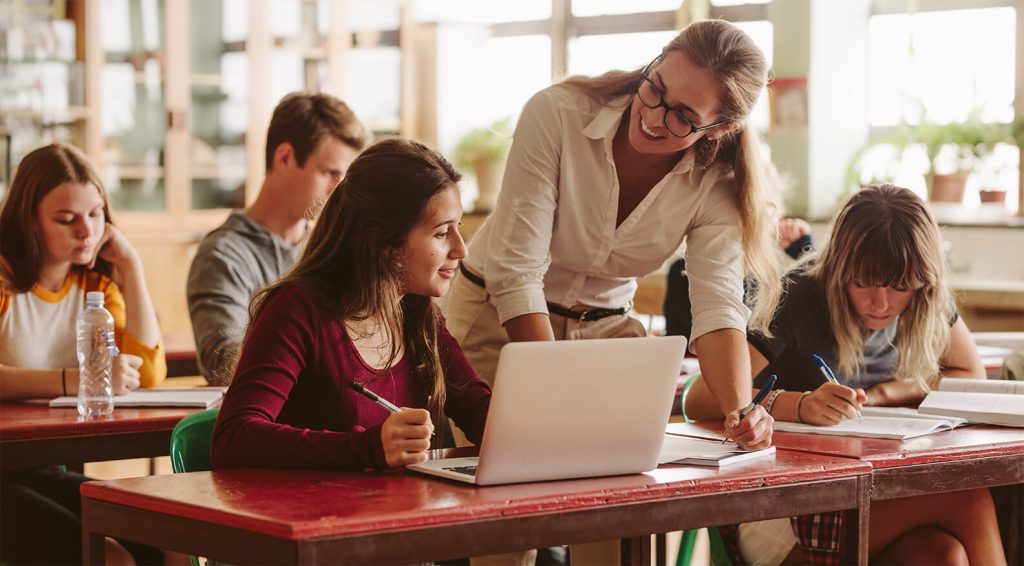 one women sitting, another women standing, the women standing is engaging the student with a laptop.